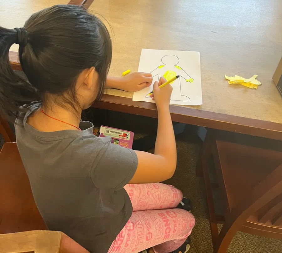 A student colors in her paper body outline at the table